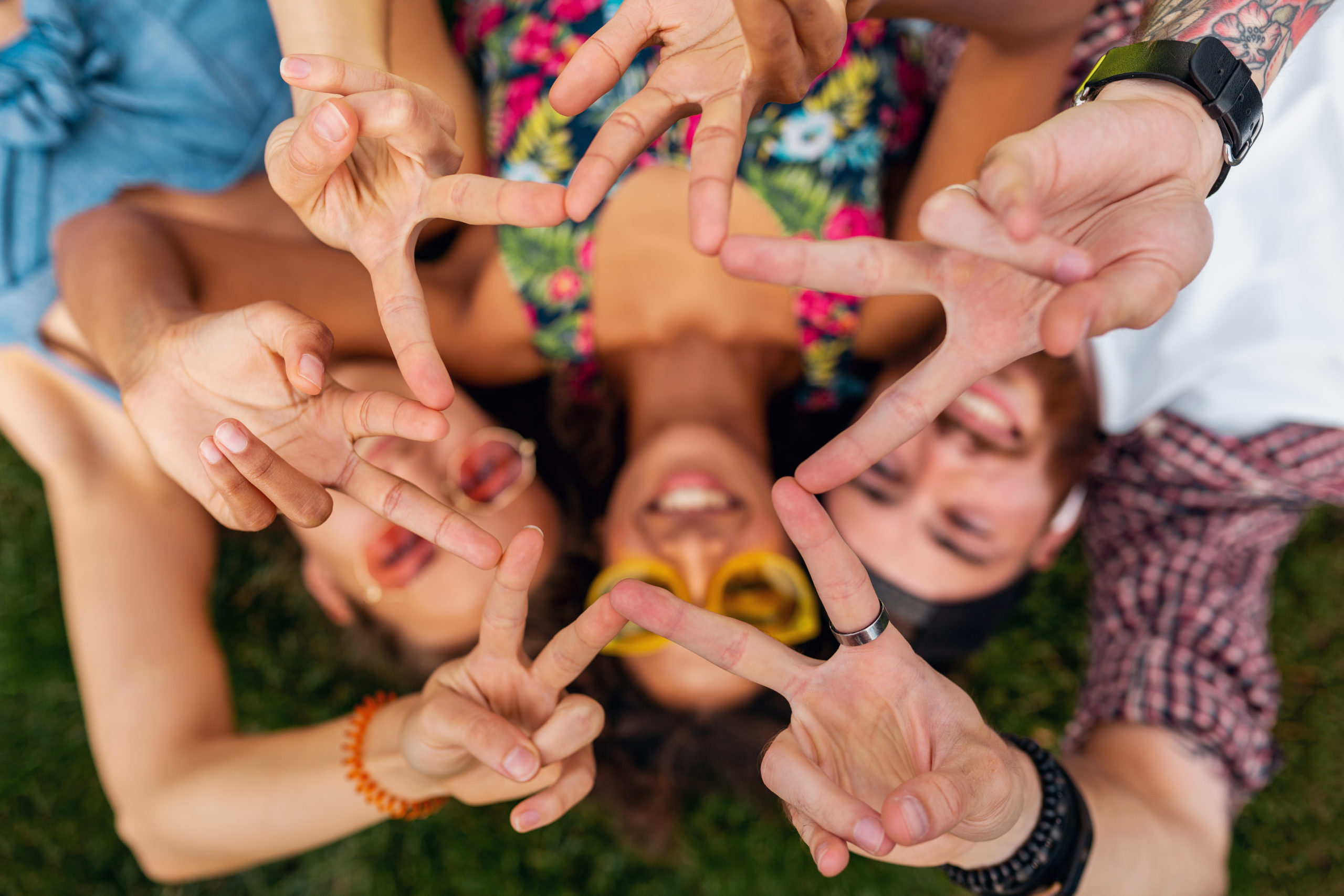 top view from above on colorful stylish happy young company of friends lying on grass in park, man and women having fun together, summer hipster fashion style, traveling smiling and showing peace sign